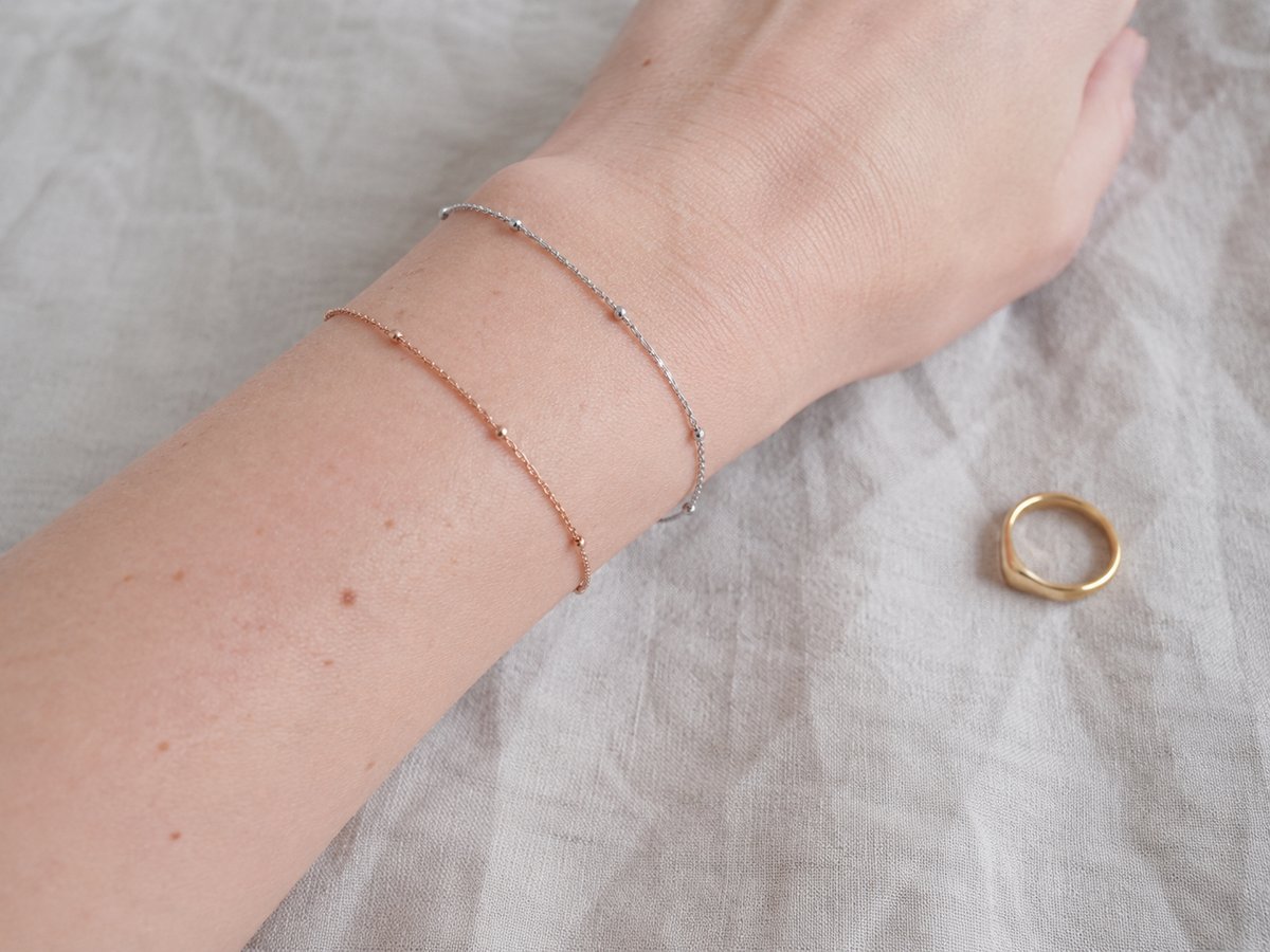 Polso con due braccialetti sottili in argento e oro rosa, con un piccolo anello dorato posato su un tessuto beige