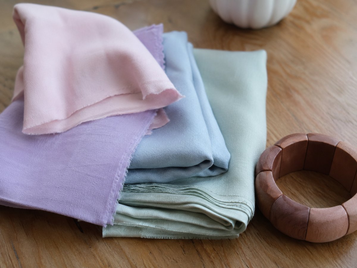 Quadrati di tessuto in toni pastello rosa, lilla, azzurro e verde salvia su un tavolo di legno con un bracciale in legno